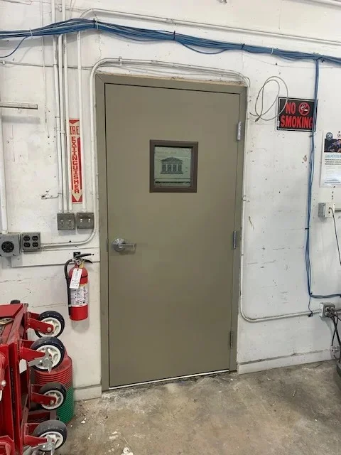 A commercial beige hollow metal door with a small square window in a workshop.