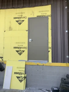A newly installed brown hollow metal door set against yellow exterior building sheathing for maintenance.