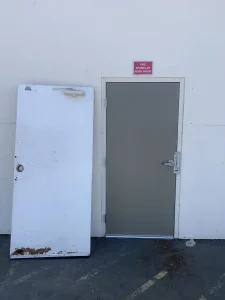 An old, rusted white door removed and placed next to a newly installed grey hollow metal door to highlight hollow metal door repair cost